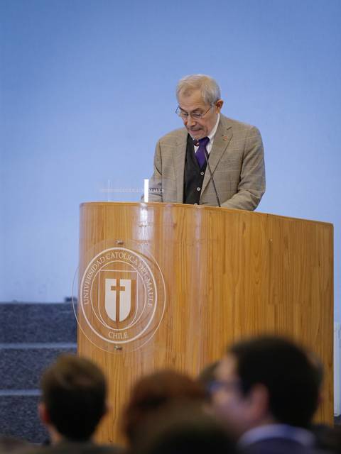 Un hombre de edad avanzada está dando una conferencia en un podio de madera en una universidad.