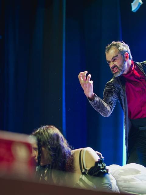 Una actuación teatral donde un hombre vestido de forma llamativa expresa una intensa emoción sobre una mujer en una escena dramática.