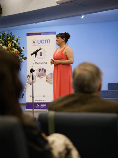 Una mujer con vestido rosa está dando un discurso frente a un micrófono en un evento relacionado con la medicina.
