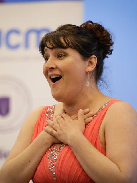 Una mujer sorprendida y emocionada, con un vestido rosa y un gesto de alegría en su rostro.