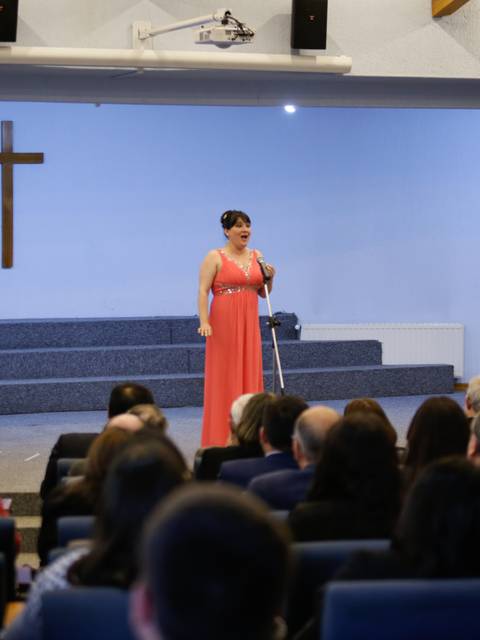 Una mujer en un vestido largo de color rosa se presenta en un auditorio con una cruz y banderas de fondo.