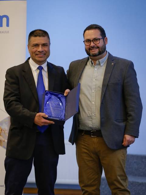 Dos hombres posan juntos sonrientes, uno sosteniendo un premio en una ceremonia.