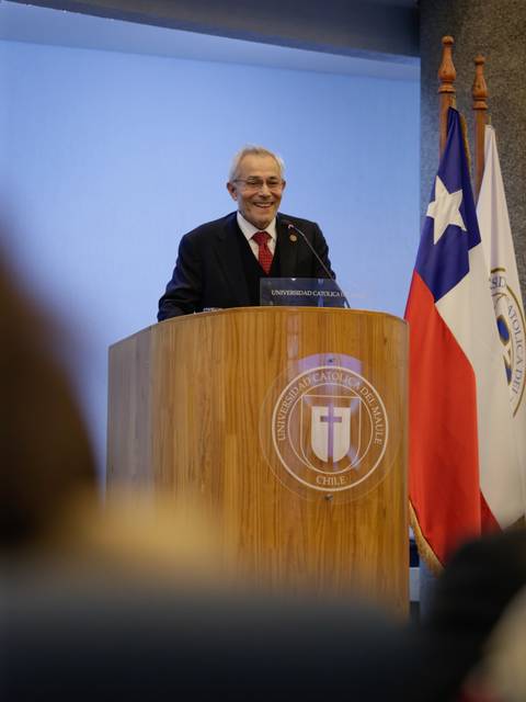 Un hombre sonriente está hablando en un podio con banderas de Chile y de una institución detrás de él.