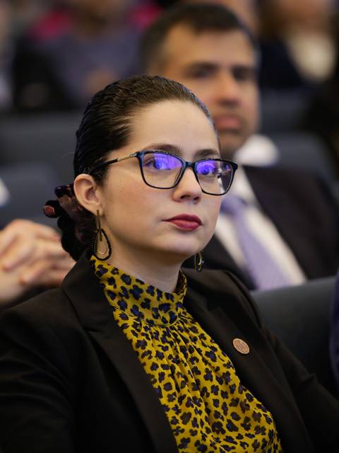 Una mujer con gafas y una blusa amarilla con estampado de leopardo escucha atentamente durante un evento en un auditorio.