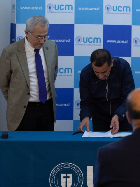 Un grupo de personas observa mientras un hombre firma documentos sobre una mesa en un evento institucional.