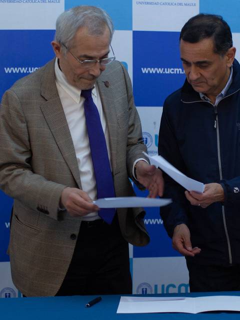 Dos hombres revisan documentos en una mesa frente a un fondo de la universidad.