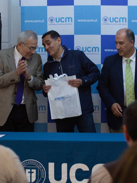 Un grupo de personas se encuentra en un evento formal donde se entrega una bolsa blanca con el logo de una institución educativa.