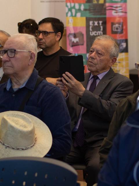 Un grupo de hombres mayores sentados en un evento, algunos usando gafas y un sombrero.