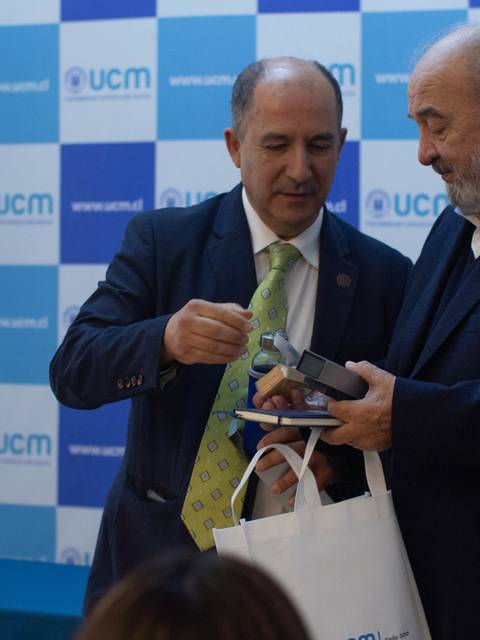 Dos hombres intercambian un objeto en un evento formal frente a un fondo con logotipos de la UCM.