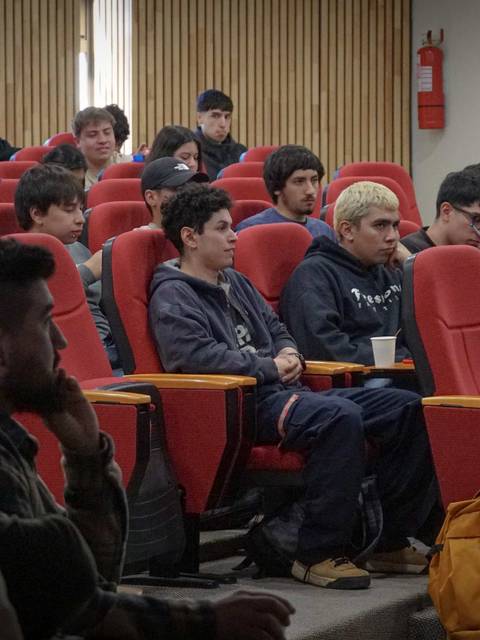 Un grupo de estudiantes sentados en un aula con asientos rojos, atentos a una presentación.