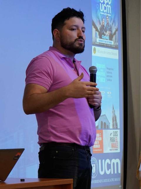 Un hombre de pie frente a un proyector, hablando mientras sostiene un micrófono.
