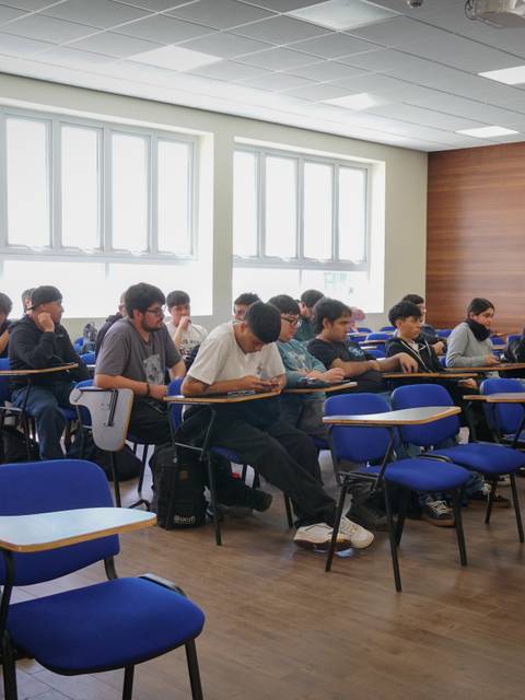 Un aula con estudiantes sentados en escritorios, algunos trabajando y otros mirando sus dispositivos.