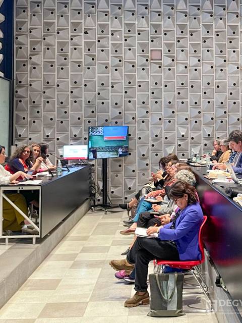 Una reunión en un salón con varias personas sentadas y presentaciones en pantalla.