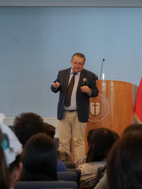 Un orador se presenta en un escenario con banderas de Chile y una institución educativa de fondo.