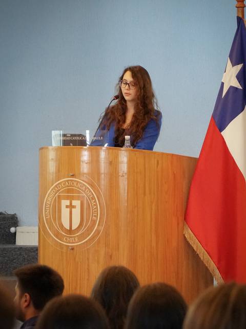 Una mujer está dando una presentación en un podio en un evento académico con una bandera chilena de fondo.