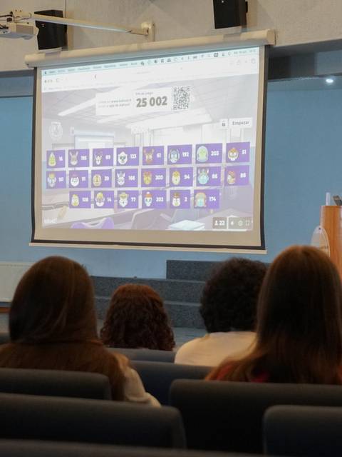 Un presentador está hablando frente a una audiencia en un auditorio con una pantalla grande detrás que muestra información.