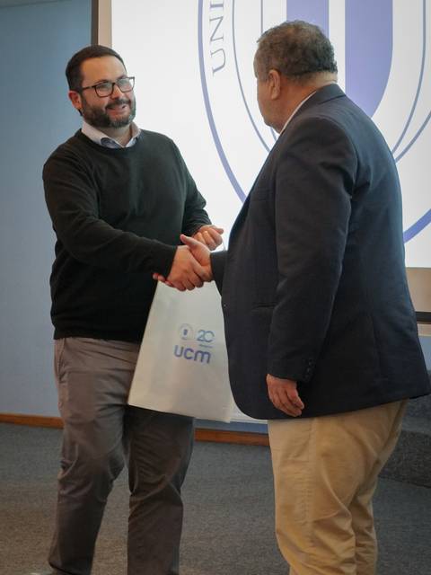 Dos hombres se estrechan la mano en un evento, uno de ellos sostiene una bolsa con el logotipo de la universidad.
