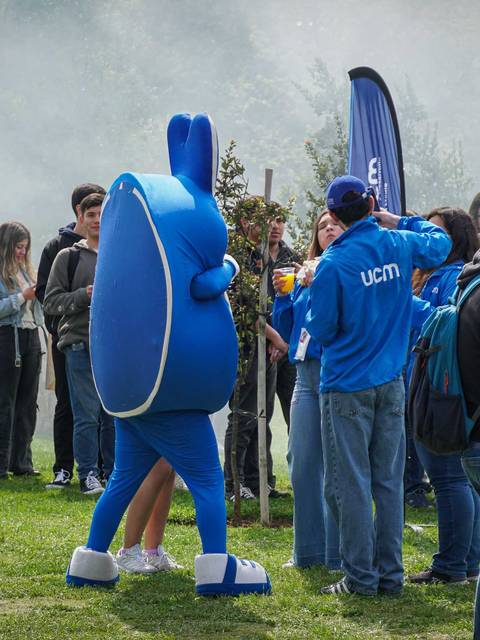 Un grupo de personas reunidas en un evento al aire libre con una mascota azul y banners de la UCM.