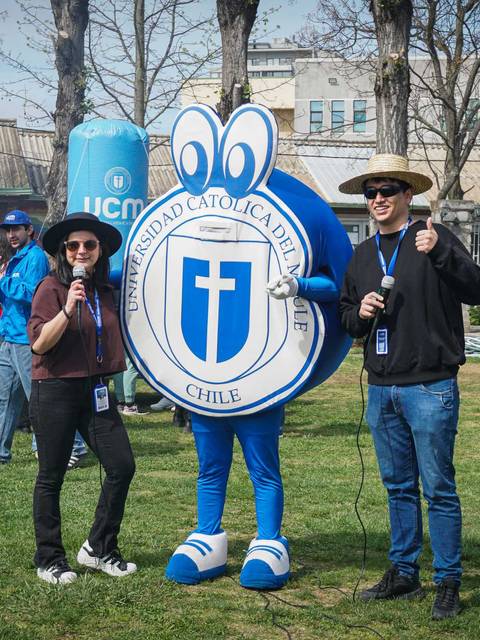 Dos personas sonrientes junto a una mascota animada en un evento al aire libre.