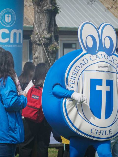 En la imagen se observa un evento con un personaje disfrazado que representa a la Universidad Católica de Chile, rodeado de estudiantes.