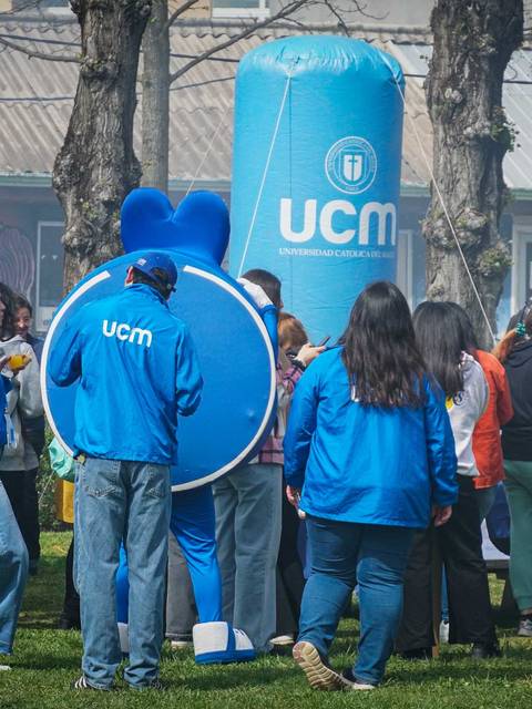 Un grupo de personas interactuando en un evento al aire libre, con un personaje disfrazado y banners de una universidad.