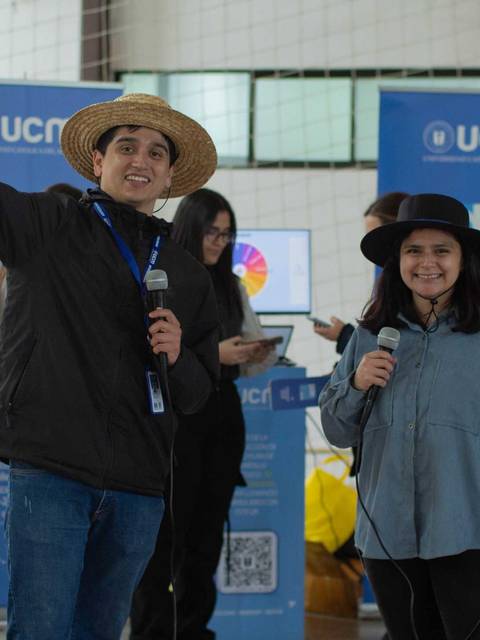 Dos personas están en un evento, una sonriendo y haciendo una señal de paz mientras sostienen micrófonos.