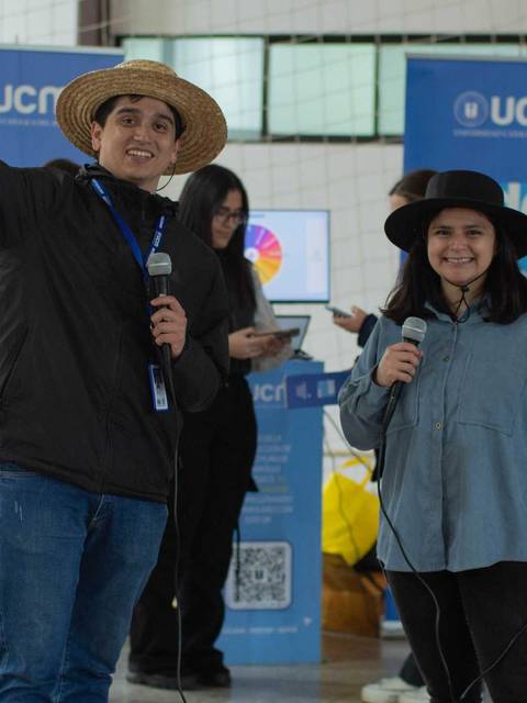 Un par de presentadores sonrientes están en un evento, uno de ellos hace un signo de paz con su mano.