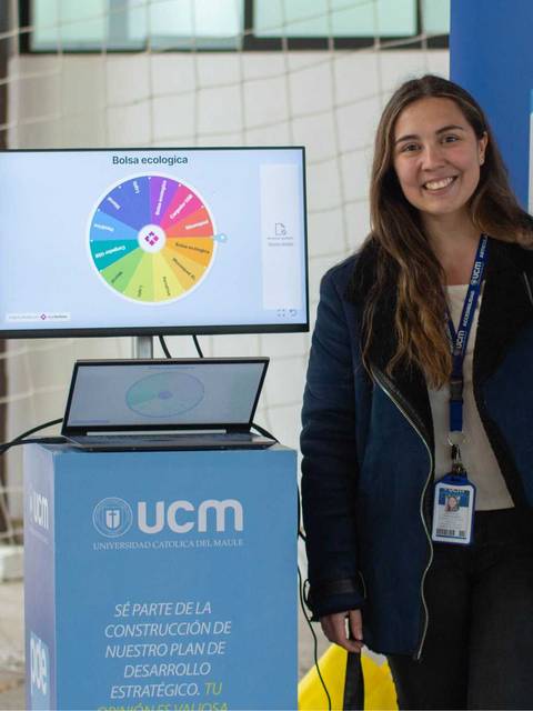 Una joven sonriente se encuentra de pie frente a una pantalla mostrando un gráfico, con banners a su alrededor que presentan información sobre un programa estratégico.