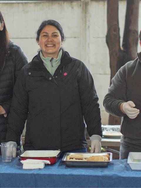 Un grupo de personas sonrientes detrás de una mesa con bebidas y comida en un evento al aire libre.