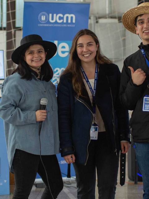 Tres jóvenes posan sonrientes en un evento, sosteniendo micrófonos frente a un stand.