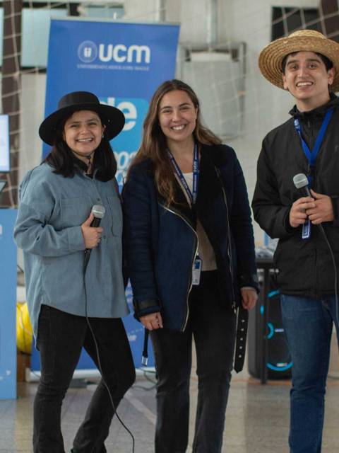 Tres personas posan sonrientes en un evento junto a pantallas y banners de la UCM.