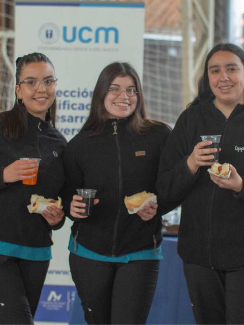 Tres jóvenes sonrientes sostienen bebidas y comida en un evento comunitario.
