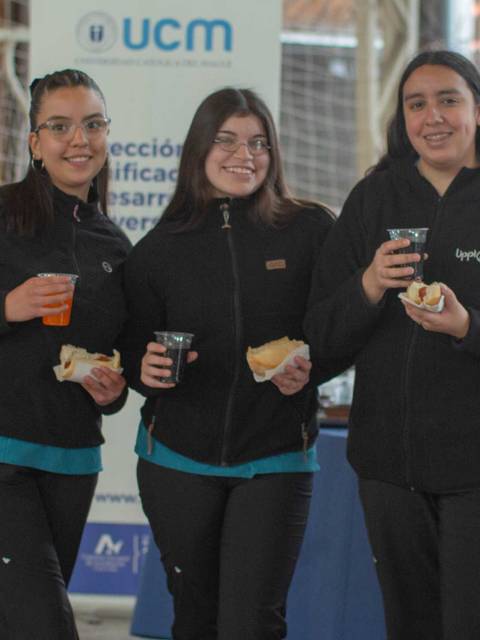 Tres jóvenes sonrientes sostienen bebidas y bocadillos en un evento escolar.