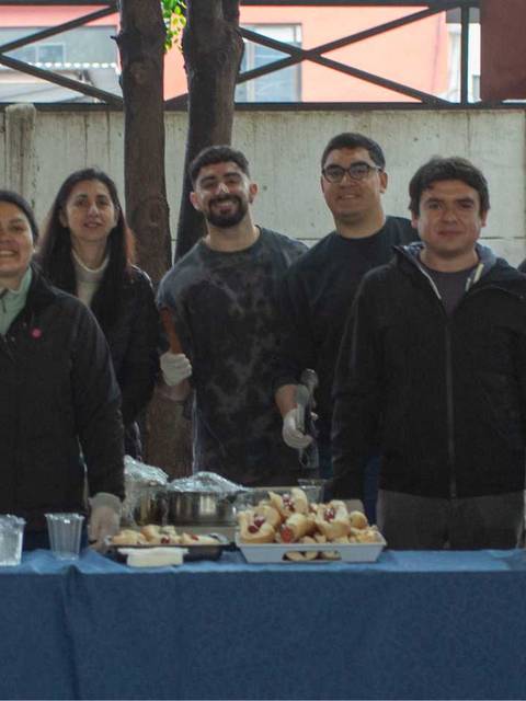 Un grupo de personas sonríe detrás de una mesa con comida y bebidas en un evento comunitario.