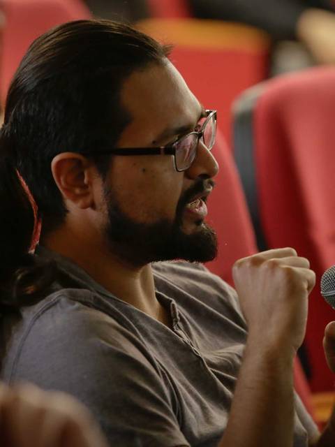 Hombre con barba y gafas sostiene un micrófono mientras participa en una conversación en un auditorio.
