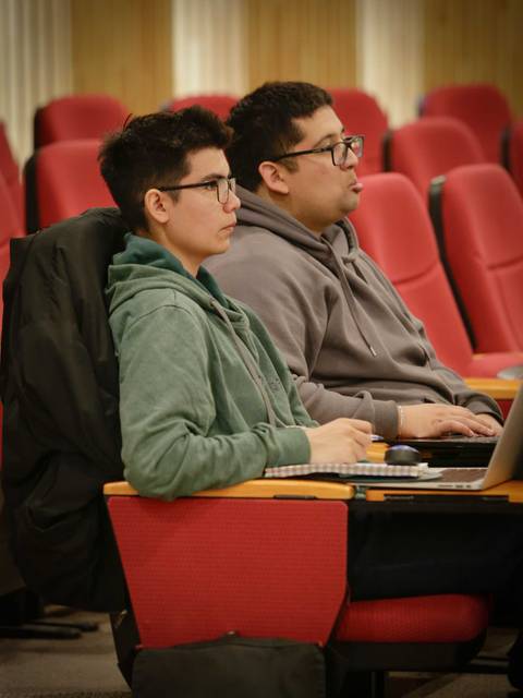 Dos personas sentadas en un aula, concentradas en sus computadoras portátiles.