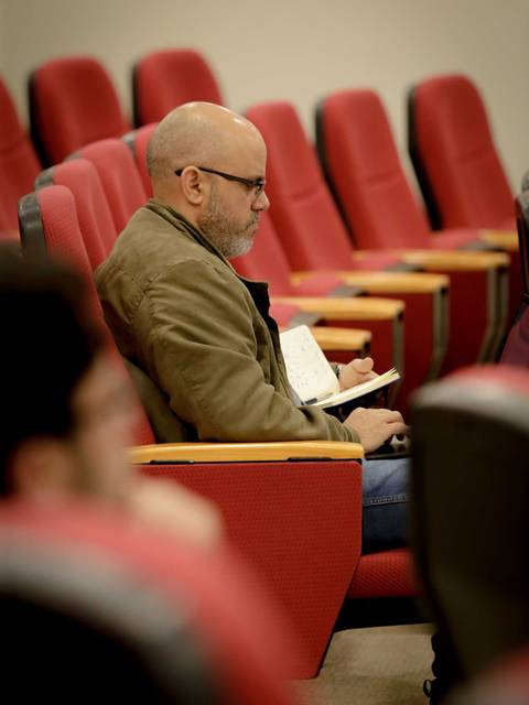 Un hombre sentado en una sala con asientos rojos, tomando notas.