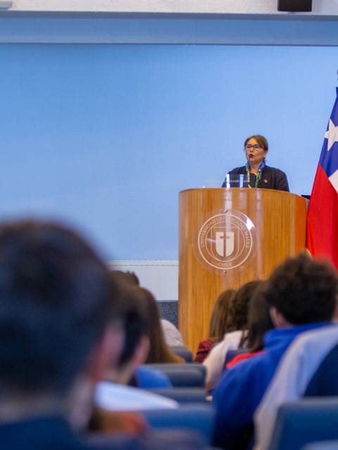 Un orador se presenta ante una audiencia en un evento con banderas chilena y de la institución en el fondo.