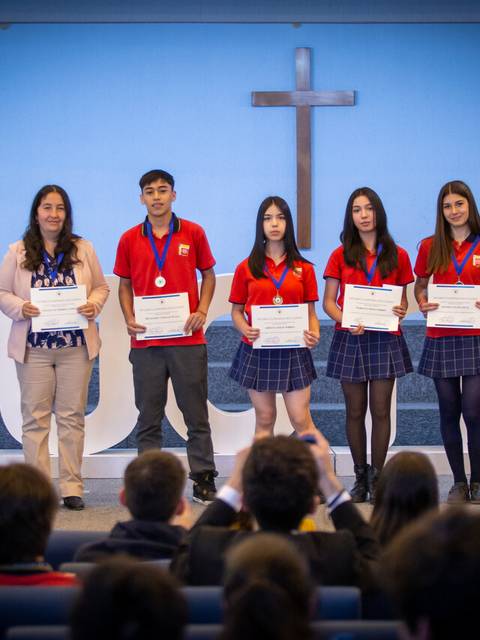 Un grupo de estudiantes recibe premios en una ceremonia escolar frente a un crucifijo.
