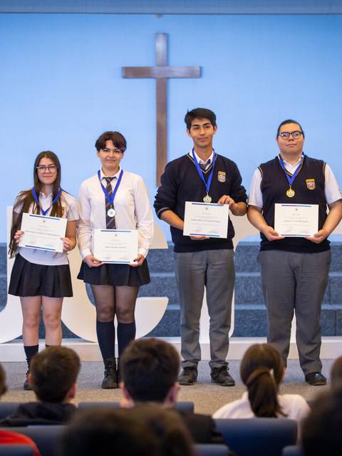 Un grupo de estudiantes recibe premios en un evento escolar frente a una cruz.