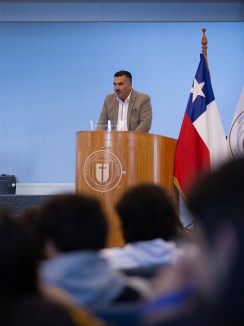Un hombre está dando una presentación en un podio frente a un público, con una bandera chilena al fondo.