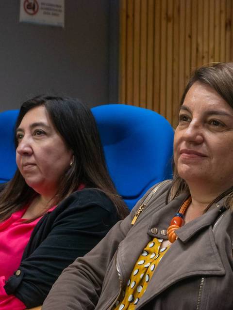 Dos mujeres sentadas en asientos azules en un espacio que parece ser una sala de conferencias.