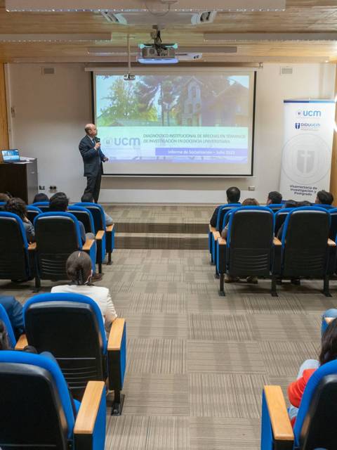 Una sala de conferencias con una presentación en pantalla y un ponente frente a la audiencia.