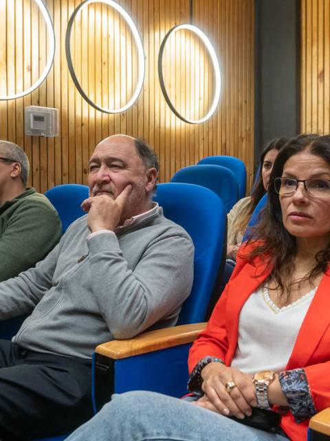 Una audiencia atenta en un salón con asientos azules y paneles de madera.