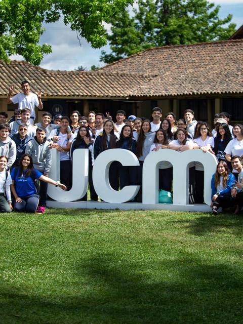 Un grupo de personas sonrientes se reúne al aire libre junto a letras grandes que forman las siglas JCM.
