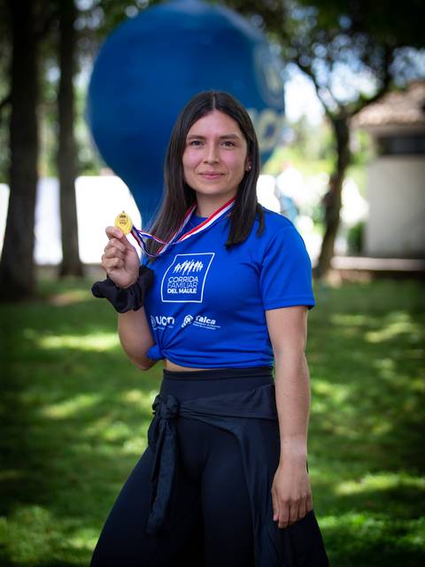 Una atleta sonriente sostiene una medalla en un evento deportivo al aire libre.
