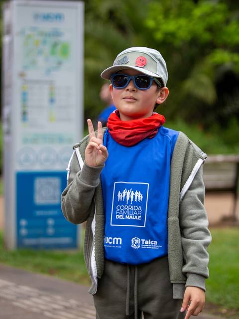 Un niño sonriente con gafas de sol y gorra, vestido con un chaleco azul, hace el símbolo de paz en un evento esportivo.