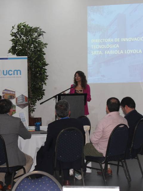 Una mujer presenta en un evento en una sala con asistentes sentados alrededor de mesas.