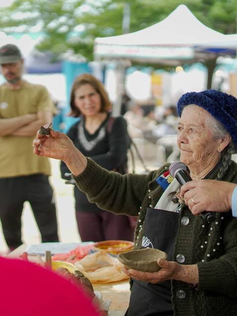 Una anciana con un sombrero azul sostiene un objeto mientras habla a un grupo de personas en un mercado al aire libre.