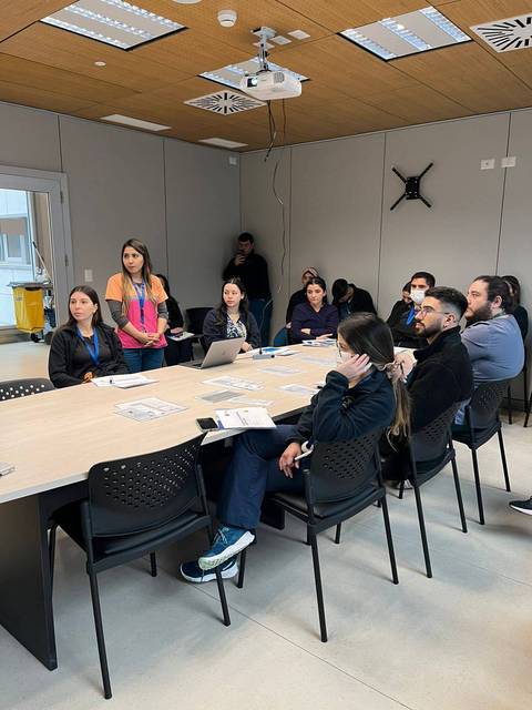 Un grupo de personas está reunido en una sala de conferencias, mientras una mujer presenta en frente del grupo.